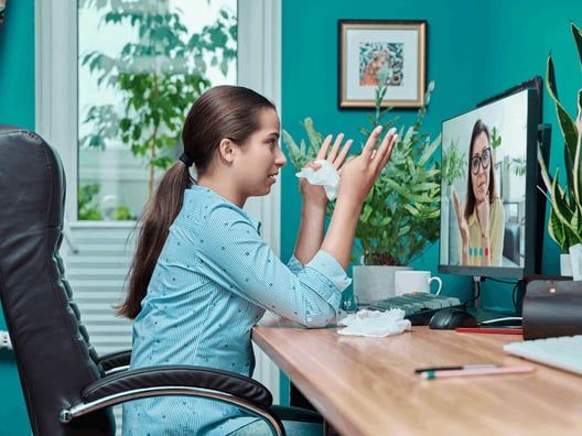 A young woman with tissue in hand, looking upset on a video call with another woman on the computer screen. They are in a home office setting.