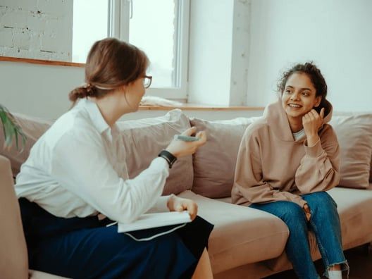 A therapist speaks with a smiling client on a sofa in a light-filled room. The therapist gestures while the client rests her chin on her hand.