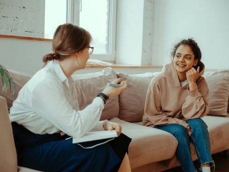 A therapist and a young woman on a sofa, talking. The woman smiles while the therapist holds a notepad, gesturing with her hand.