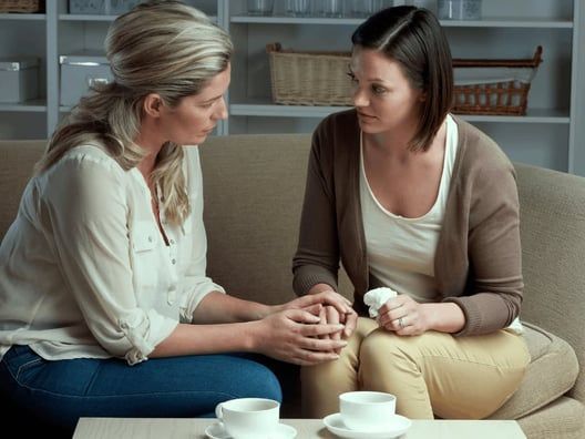 Two women sit on a couch, one holding the other's hand, looking concerned. Softly lit living room, coffee cups visible.