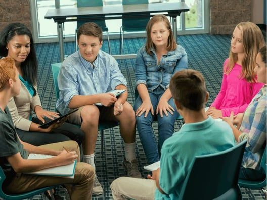 A group of diverse teens sits in a circle in a room with blue carpet and a window, engaging in a discussion.