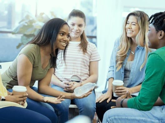 A diverse group of women sitting in a circle, smiling and talking, some holding coffee cups, in a bright room.