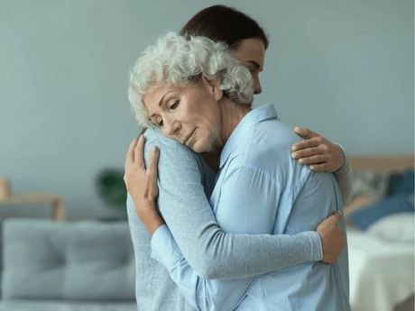 A younger person hugs an older woman, both appearing saddened, indoors. The woman has gray hair, and they are embracing in front of a blurred background.