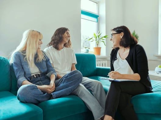 A therapist in a black jacket consults with a couple on a teal couch. The couple is smiling at each other.