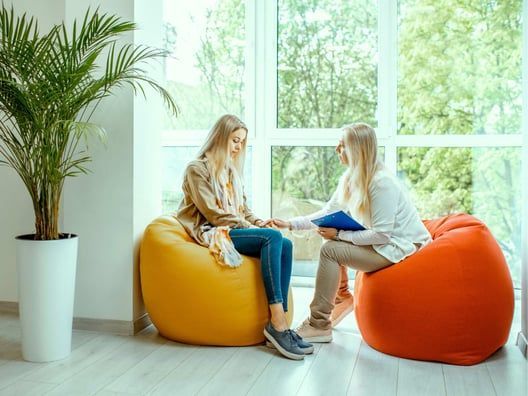 A therapist comforts a client sitting on an orange beanbag chair, holding her hand, indoors by a window with a plant.