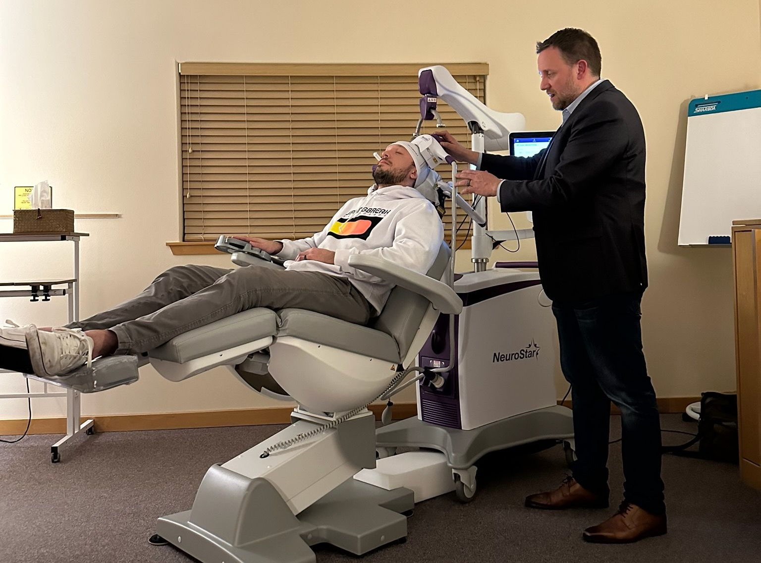 A doctor in a white coat reviews a tablet with a seated patient in an exam room. Both are focused on the tablet.