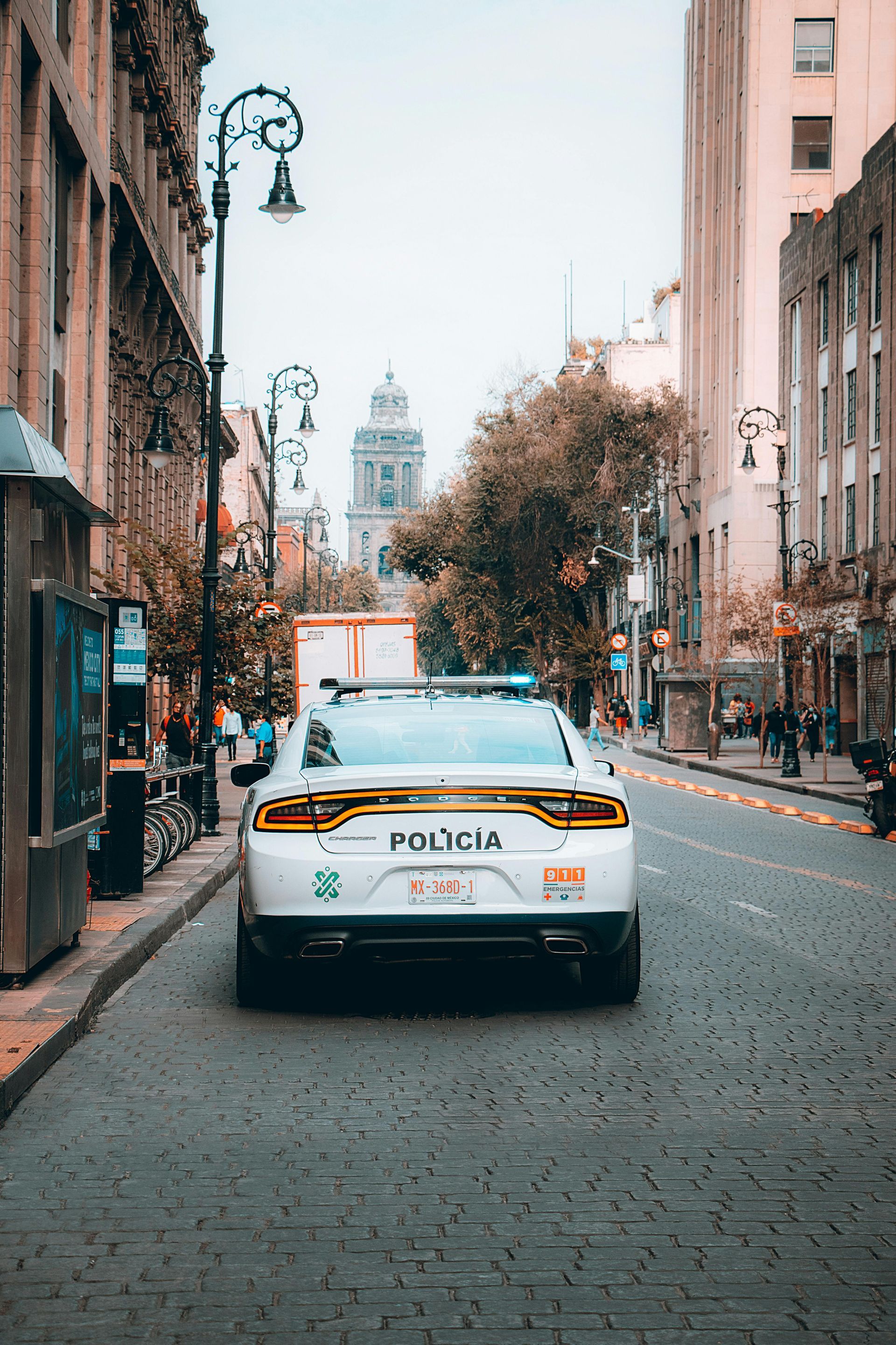 A white police car parked in the middle of a cobblestone street in a city, facing a distant building between tall halls.