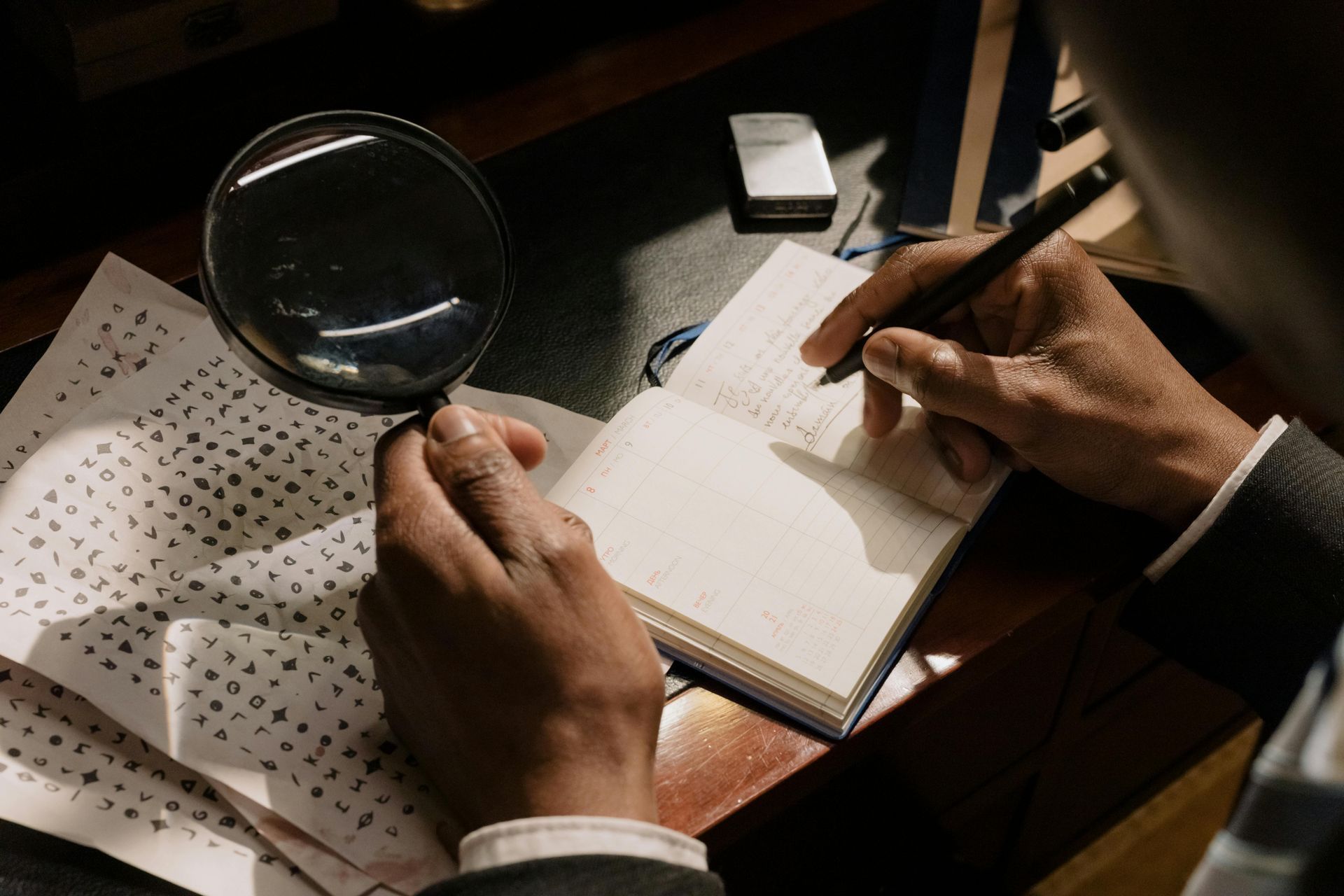 Person using a magnifying glass to examine a document and taking notes in a journal.