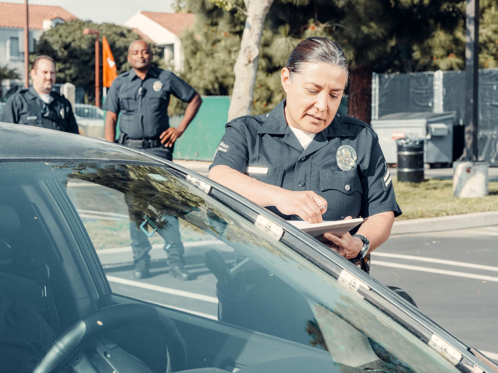 A police officer writes on a clipboard next to a car, while two other officers stand in the background.