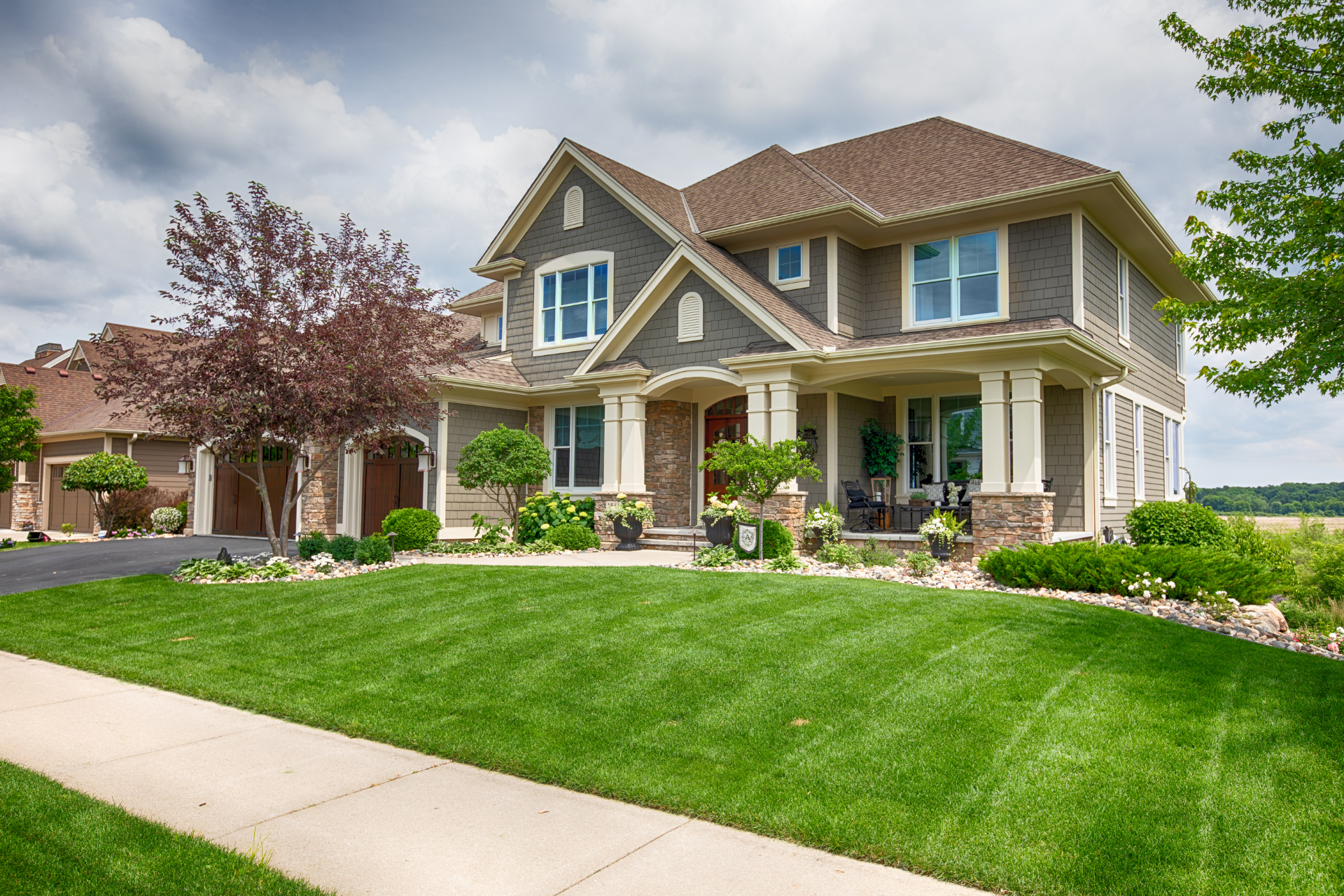 Two-story suburban house with a manicured green lawn, front porch, and trees in the yard.