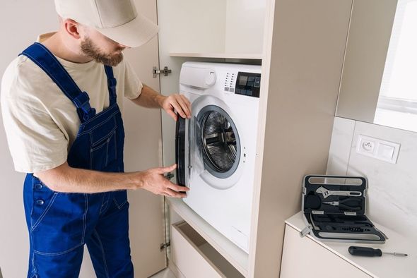 A person in overalls fixing a washing machine in a cabinet, with a toolbox nearby.
