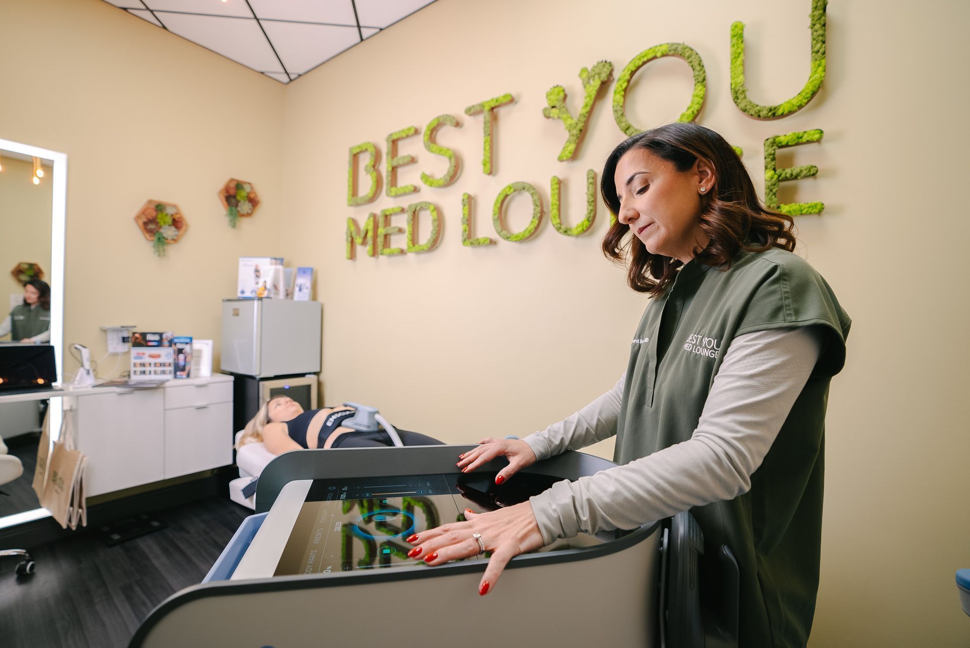 Woman in clinic examines patient on treatment bed, “Best You Med Lounge” sign visible.