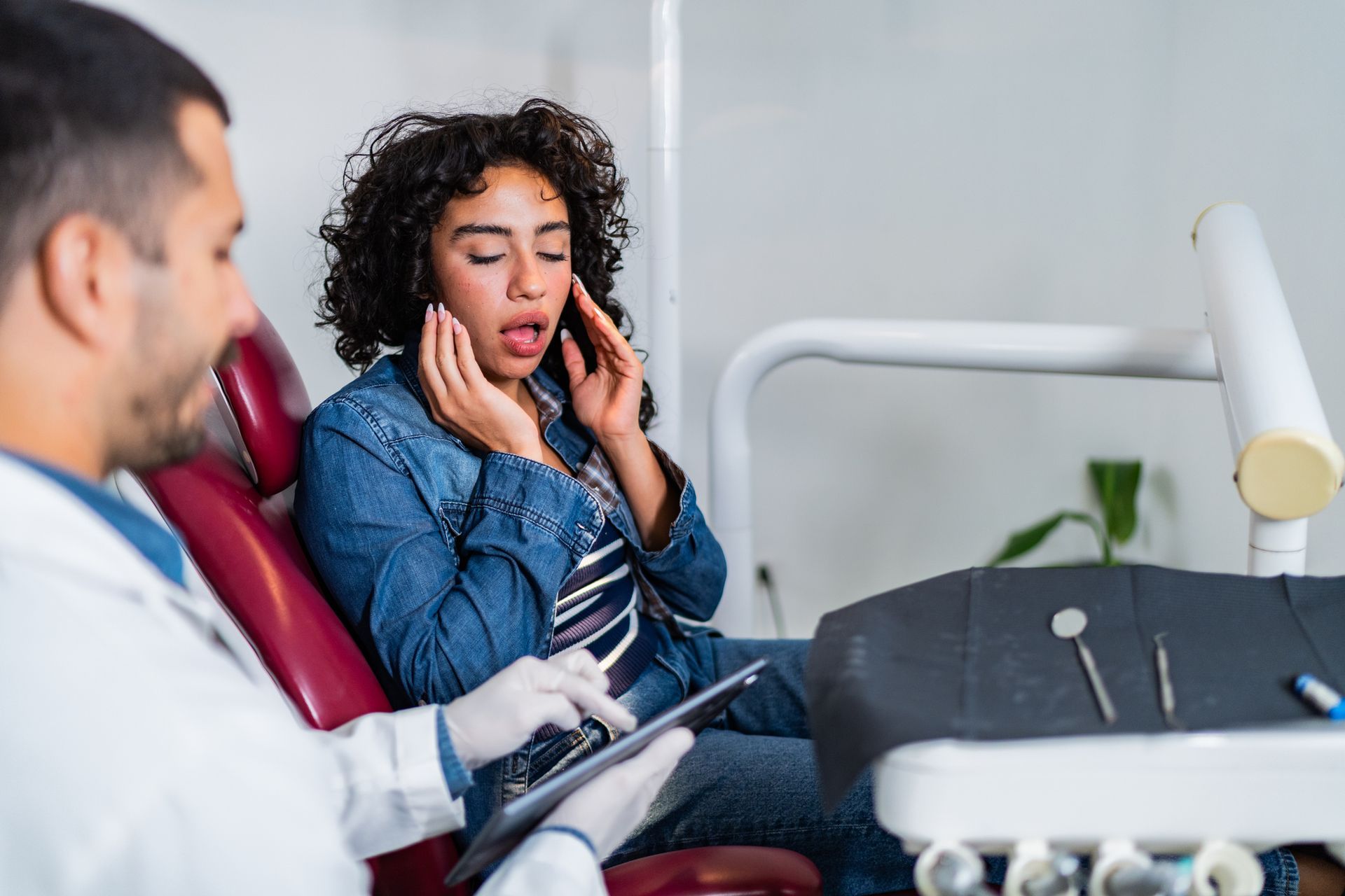 Dentist examines patient's mouth in dental chair. Patient looks distressed, holding face.