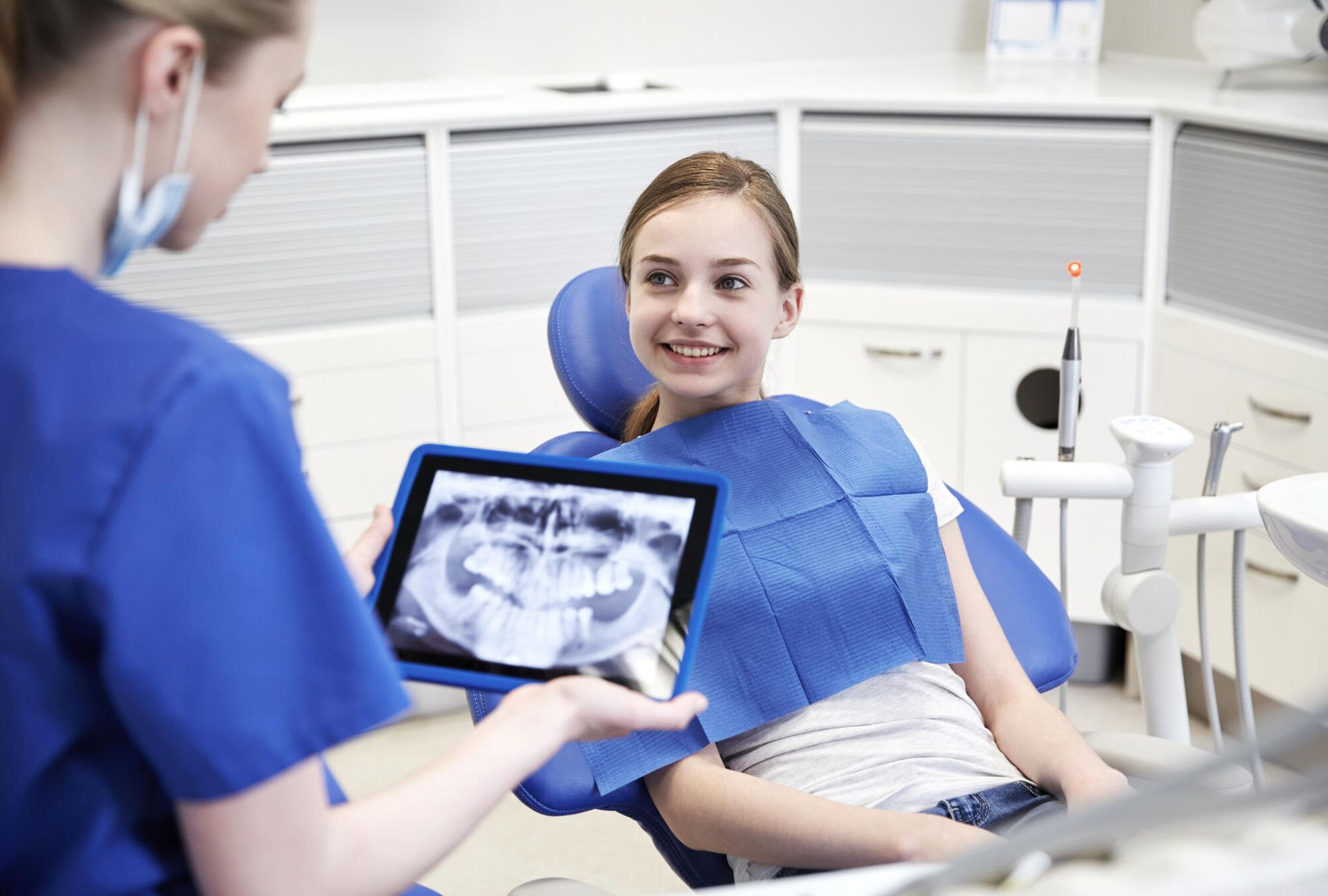 A young girl is sitting in a dental chair while a dentist shows her an x-ray of her teeth.