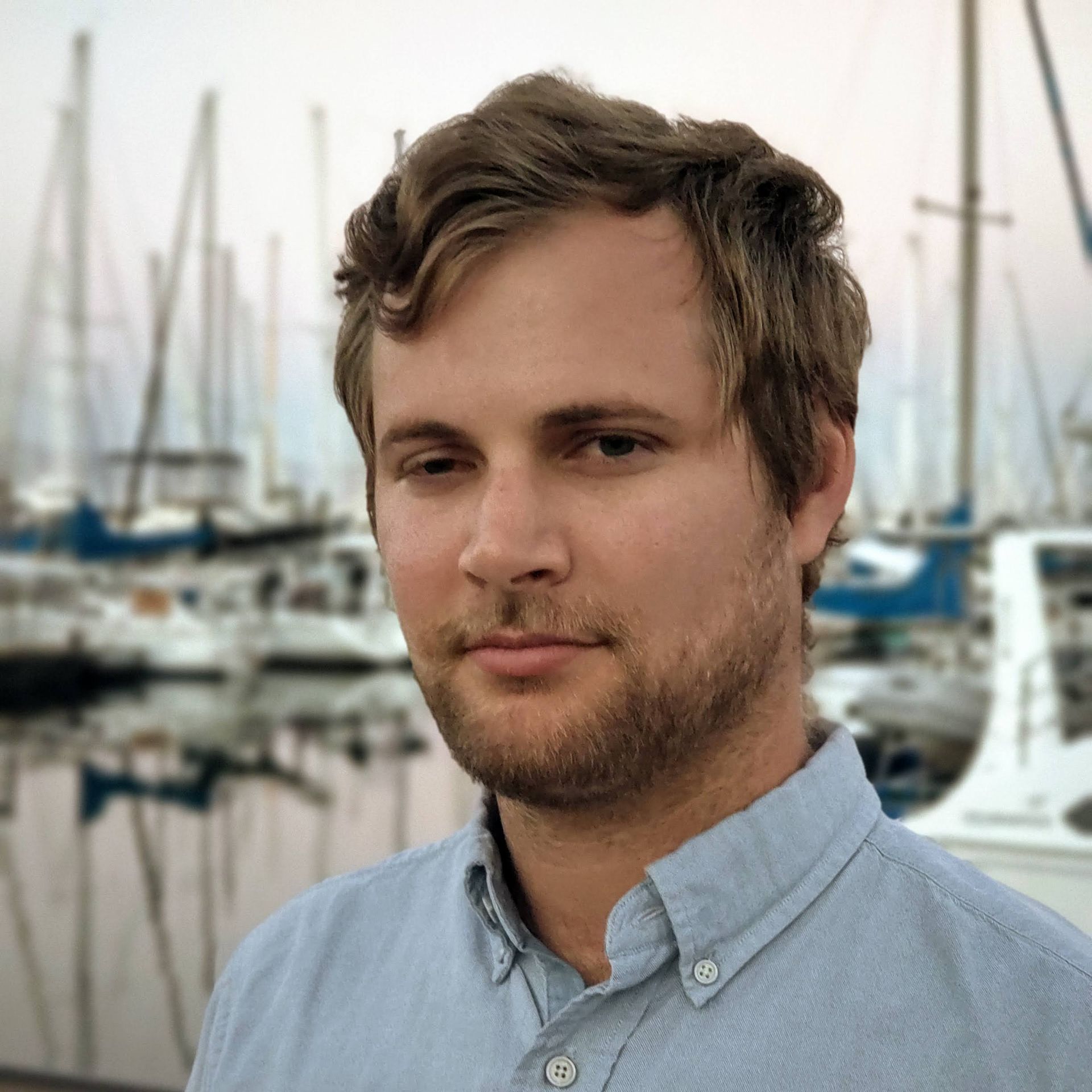 Man with light brown hair and beard in front of sailboats. Wearing a blue button-down shirt.