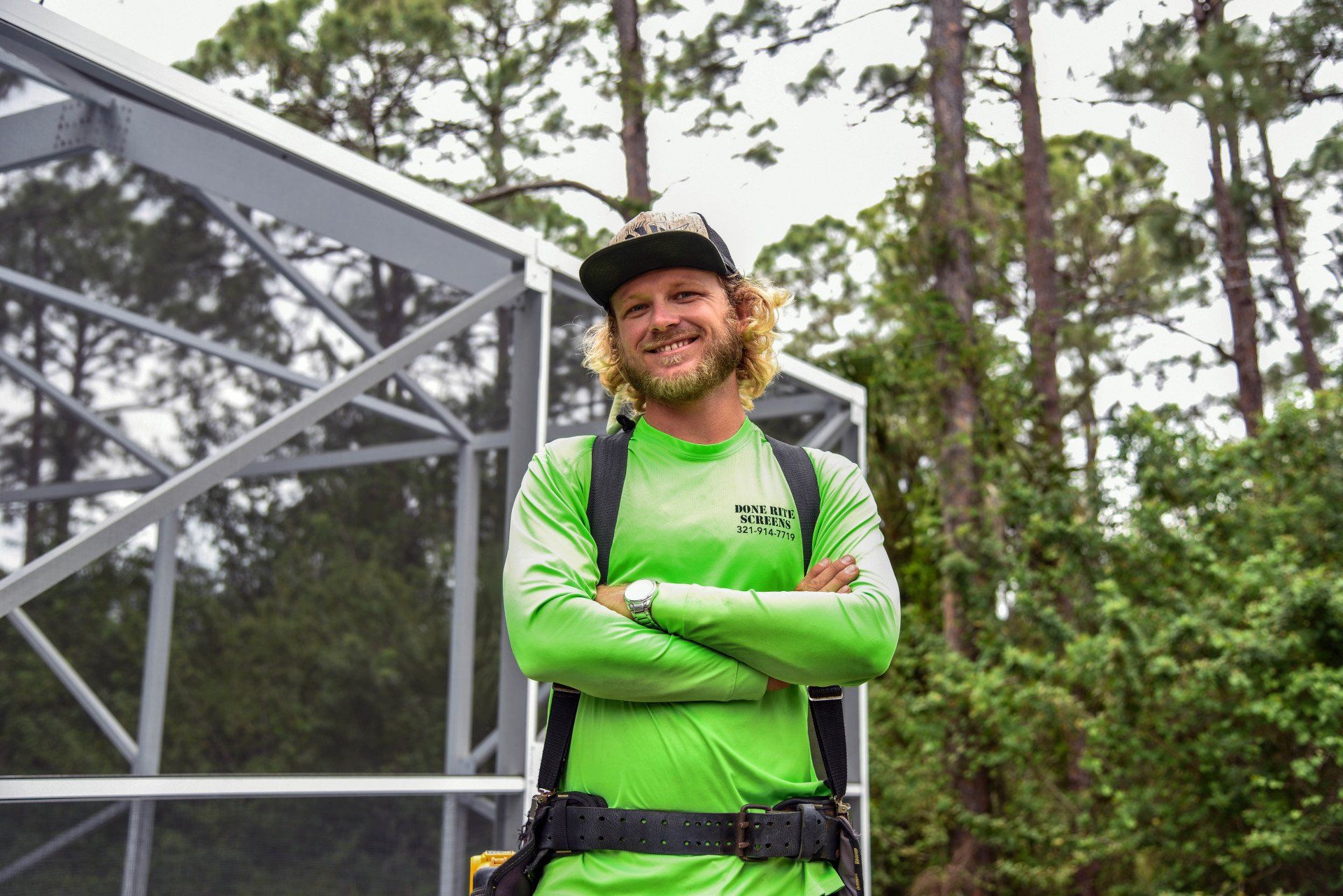 A man in a green shirt and hat is standing in front of a greenhouse.