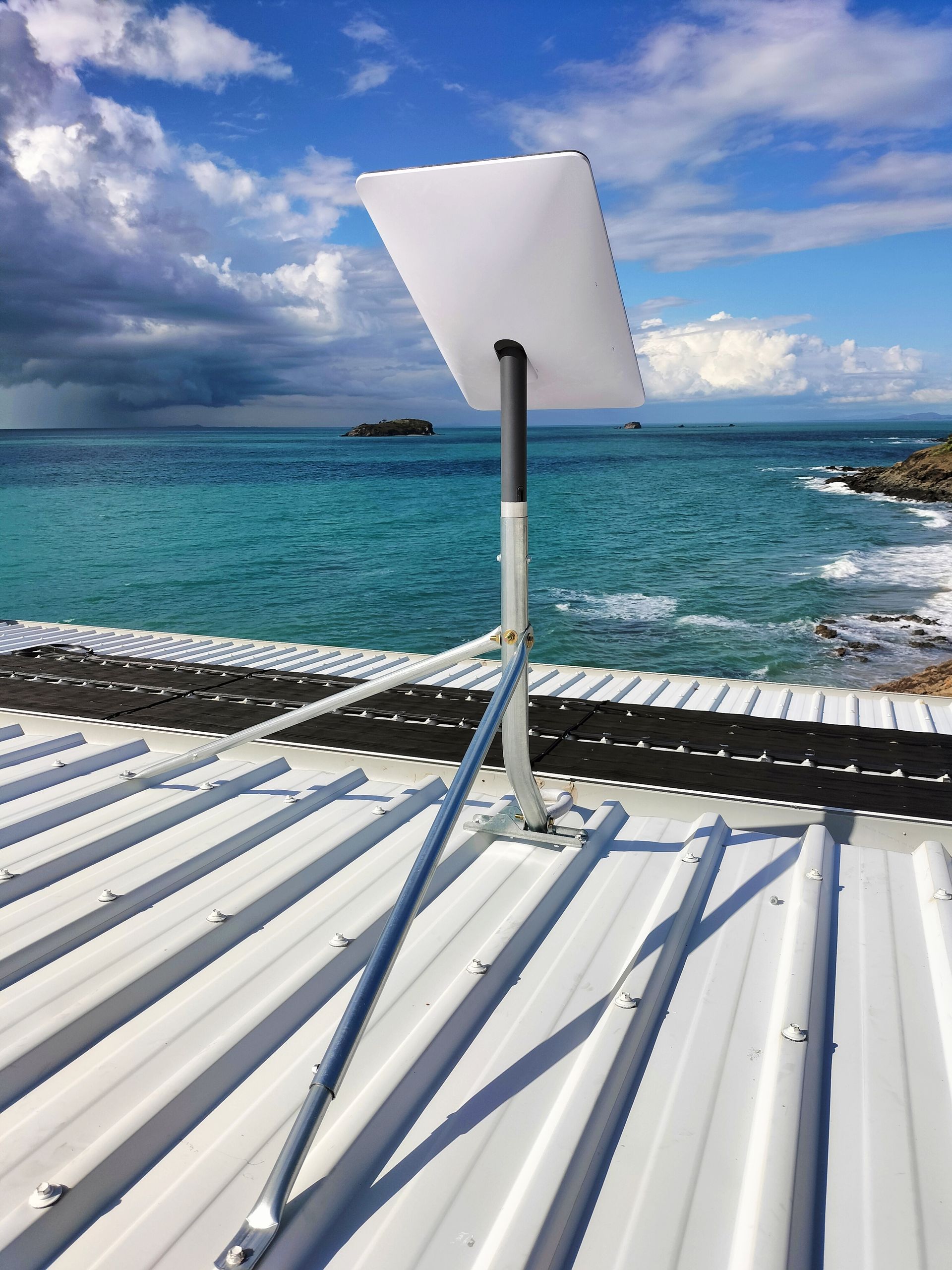 Satellite dish mounted on a corrugated metal roof, with ocean and cloudy sky in the background.