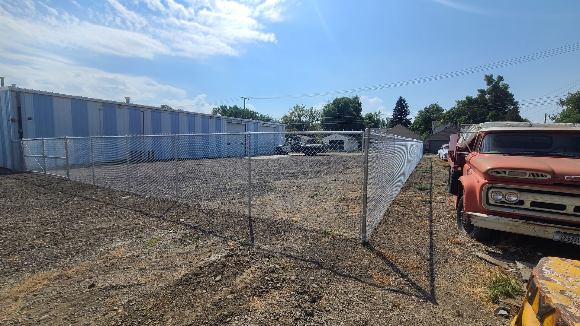 A row of old trucks are parked in a gravel lot behind a chain link fence.