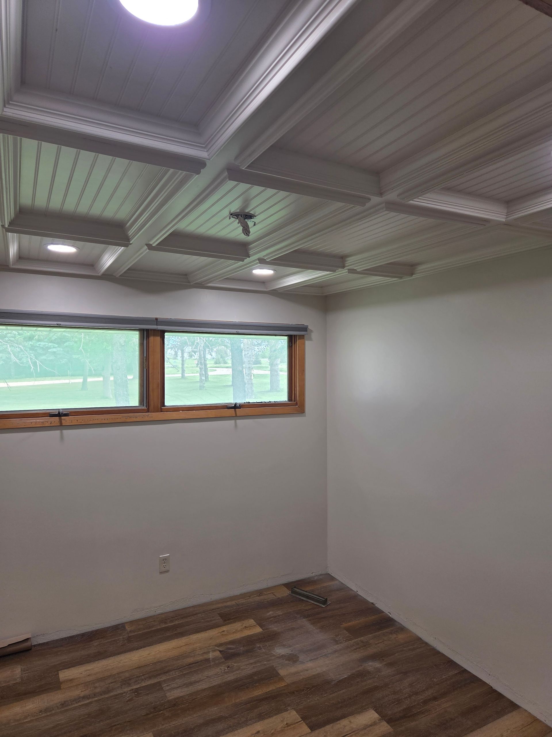Empty room with white walls, wood-look floor, and a small window under a coffered ceiling.
