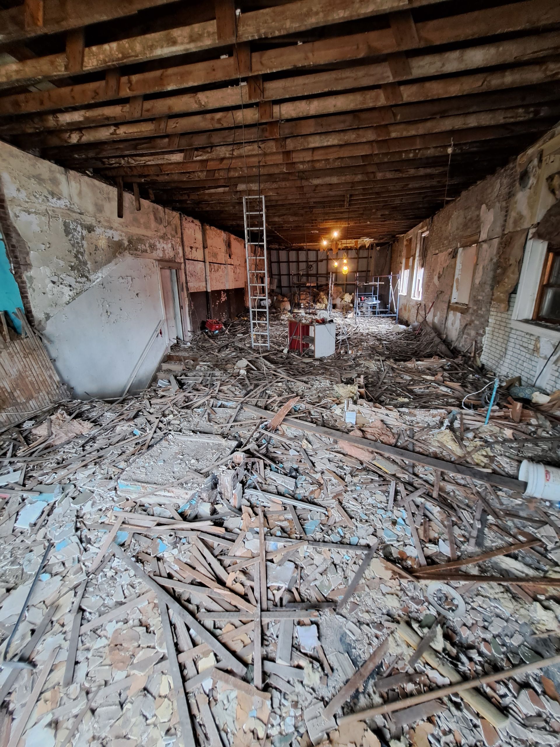 Collapsed building interior with rubble-strewn floor, exposed beams, and a ladder in the center.