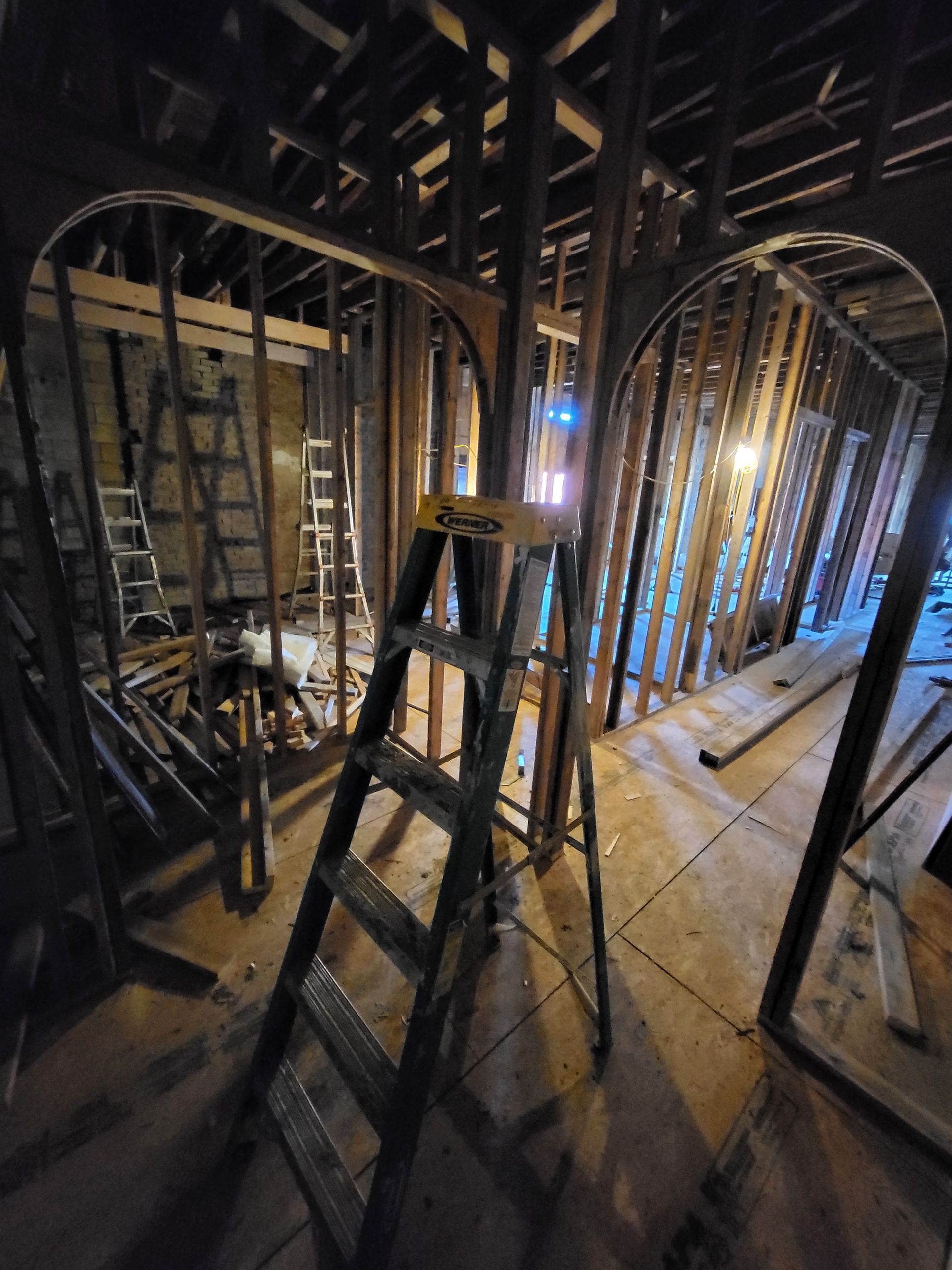 Dim attic under renovation with ladders, exposed beams, wiring, and slanted roof framing.