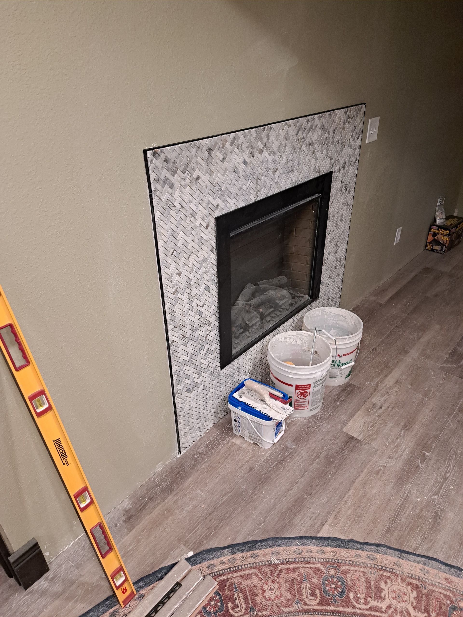 Unfinished fireplace with white tile surround, paint buckets, and a level leaning against a beige wall.
