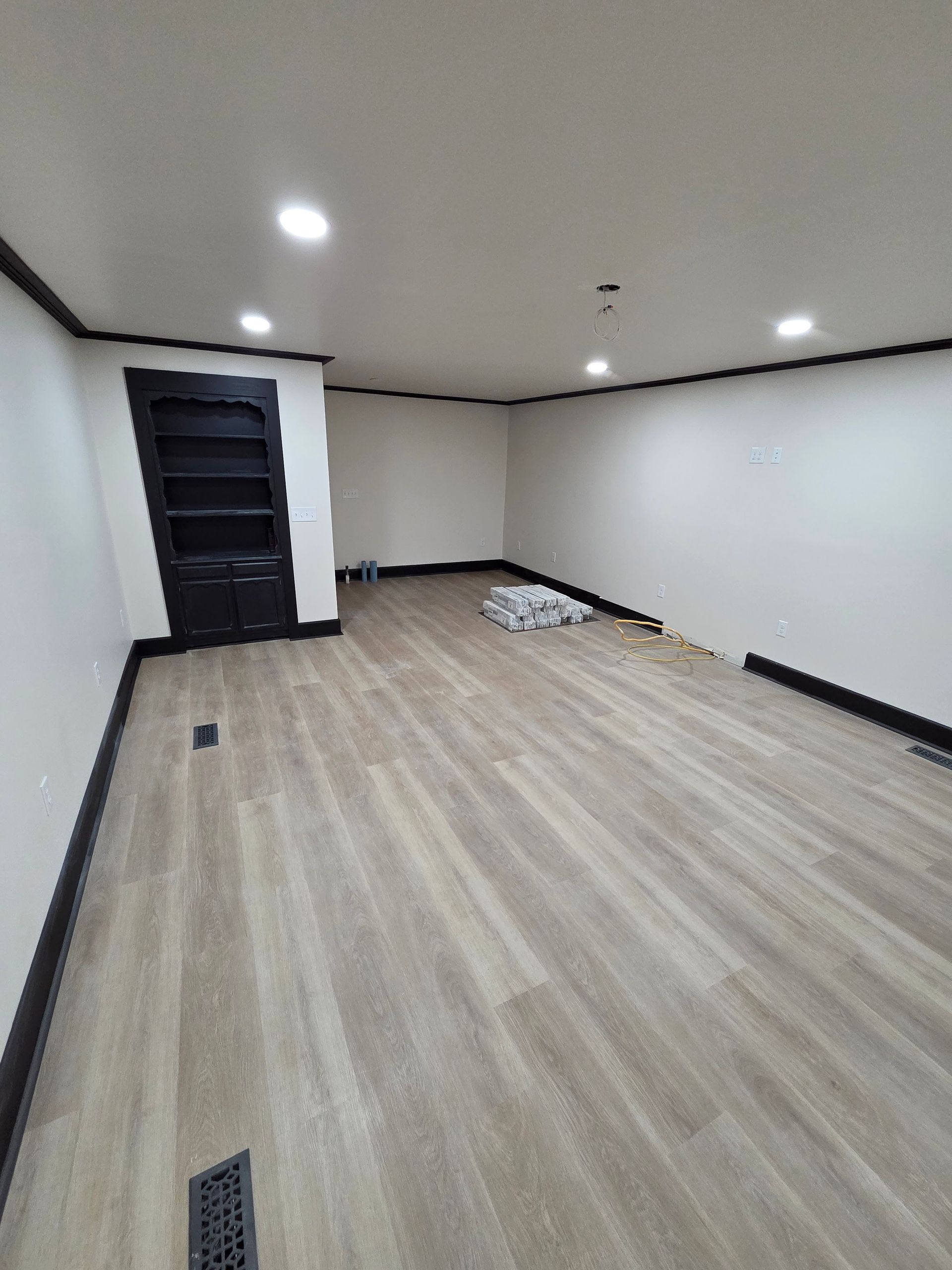 Empty modern room with light wood flooring, white walls, recessed lights, and a dark door.