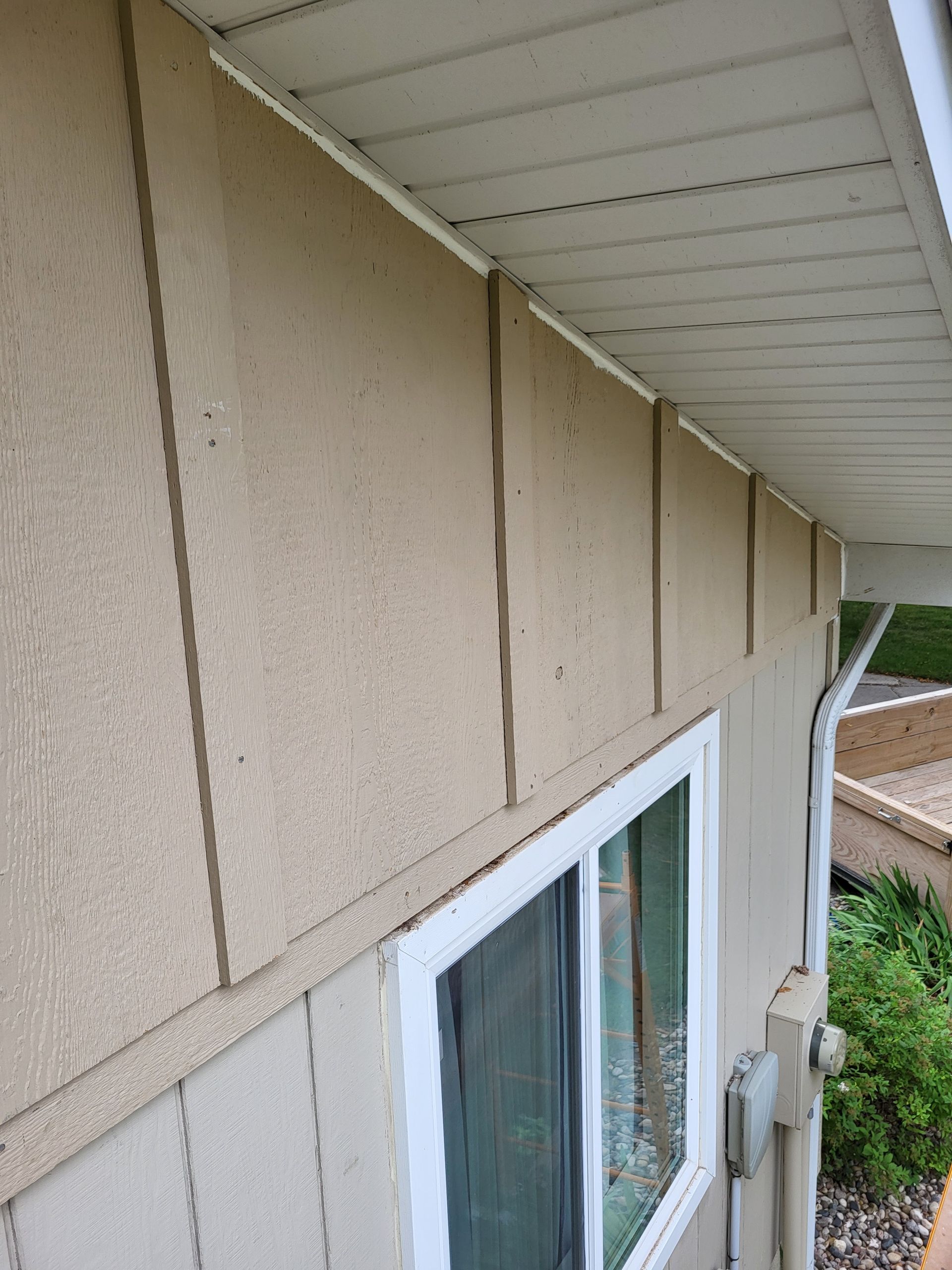 Side of a beige house with vertical siding and a white-framed window under the eaves.