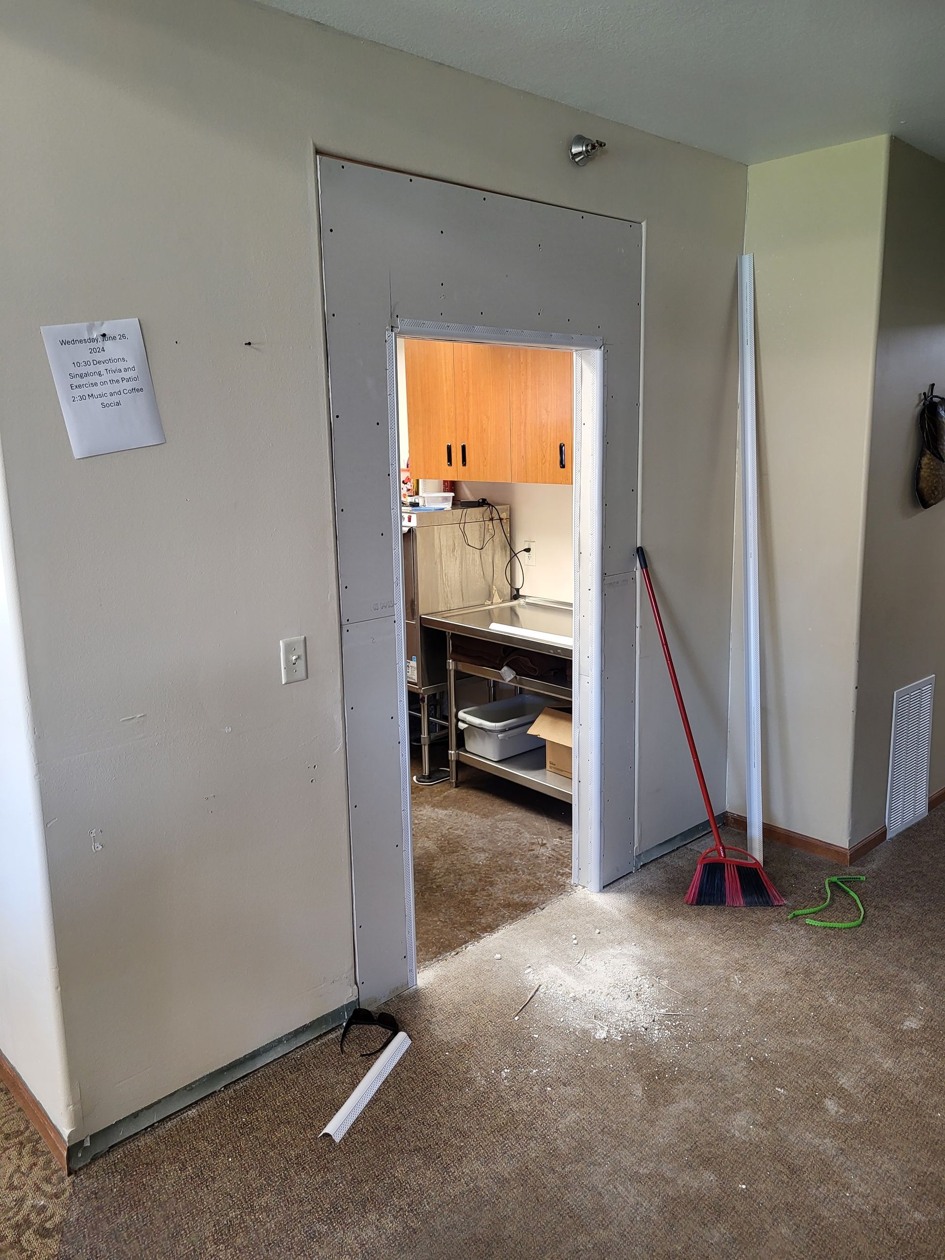 Doorway into a kitchen during renovation, with tools and a broom on a carpeted floor.