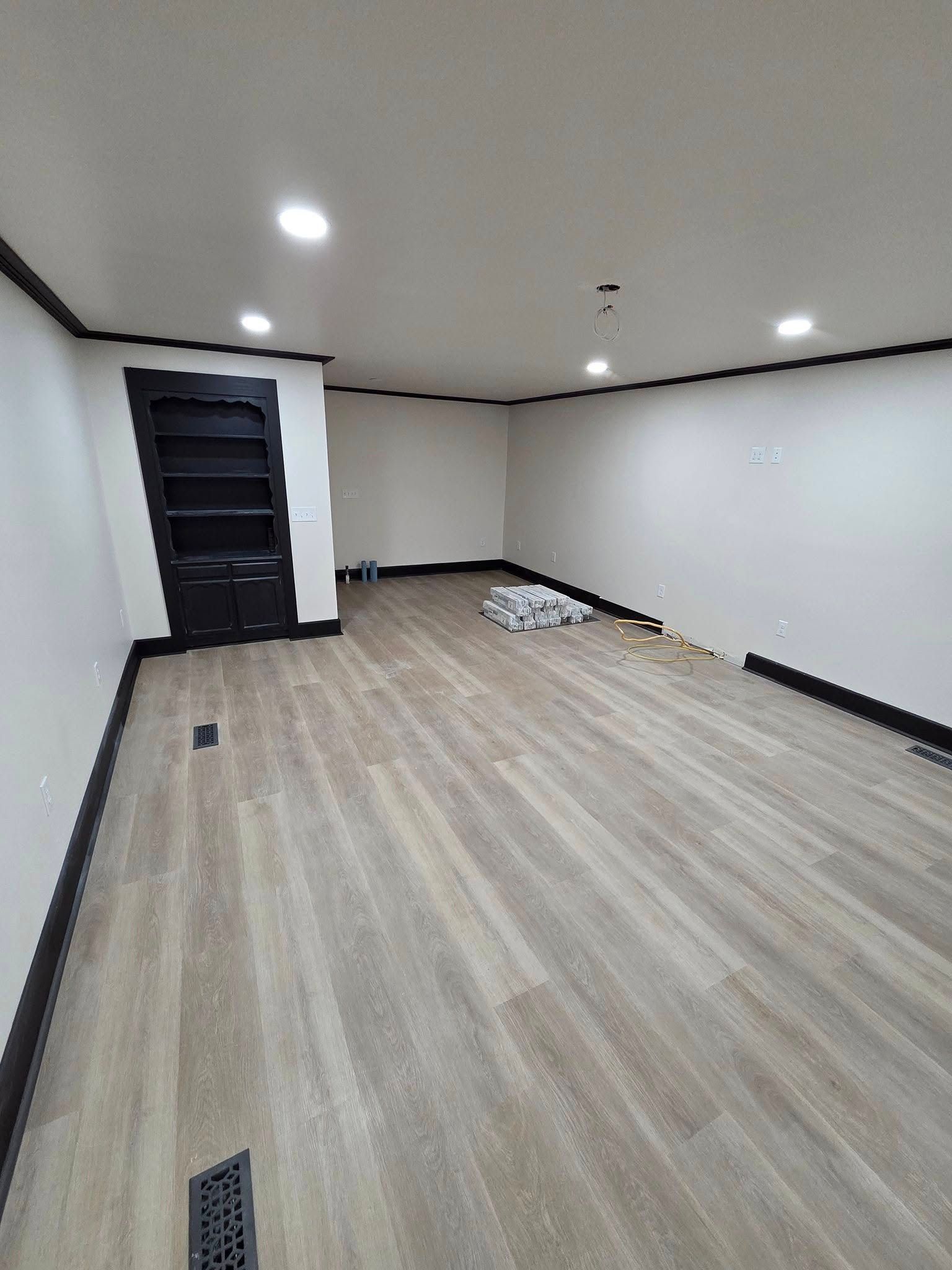 Empty room with light wood floors, white walls, recessed ceiling lights, and a dark door on the left