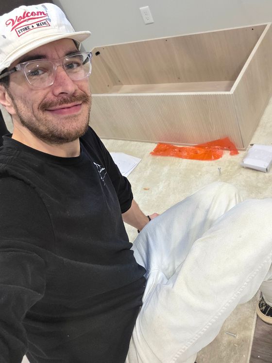 Person smiling while sitting beside a bed or couch with white bedding and an orange package nearby.