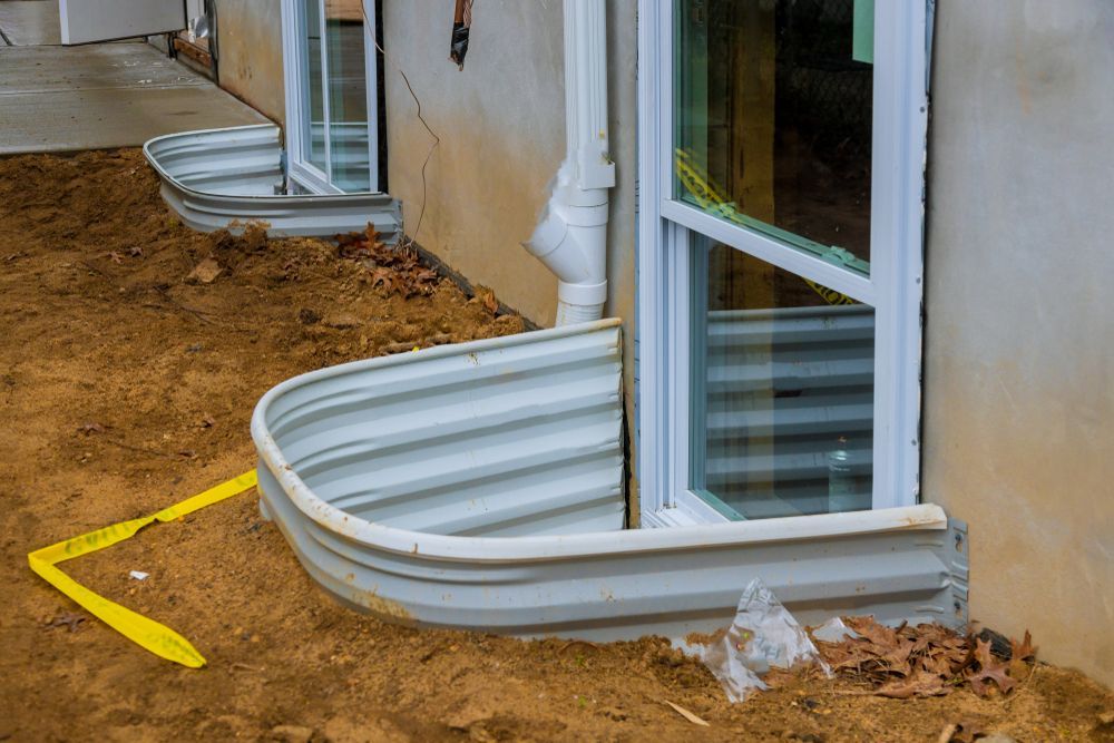 Basement window well with corrugated metal cover beside a house wall and muddy ground