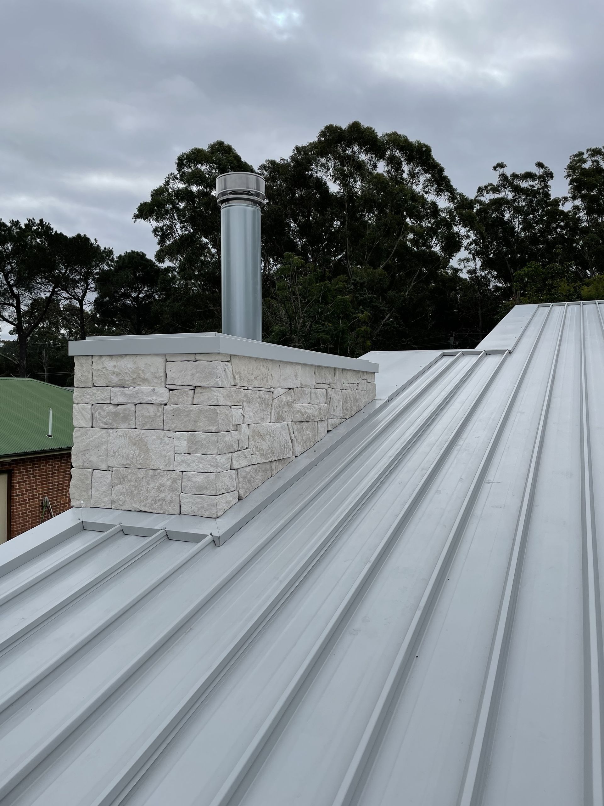 Chimney on a metal roof, gray brick base, silver flue, overcast sky, trees in the background. — R&M Sheetmetal Nowra Metal Roofing in South Nowra, NSW