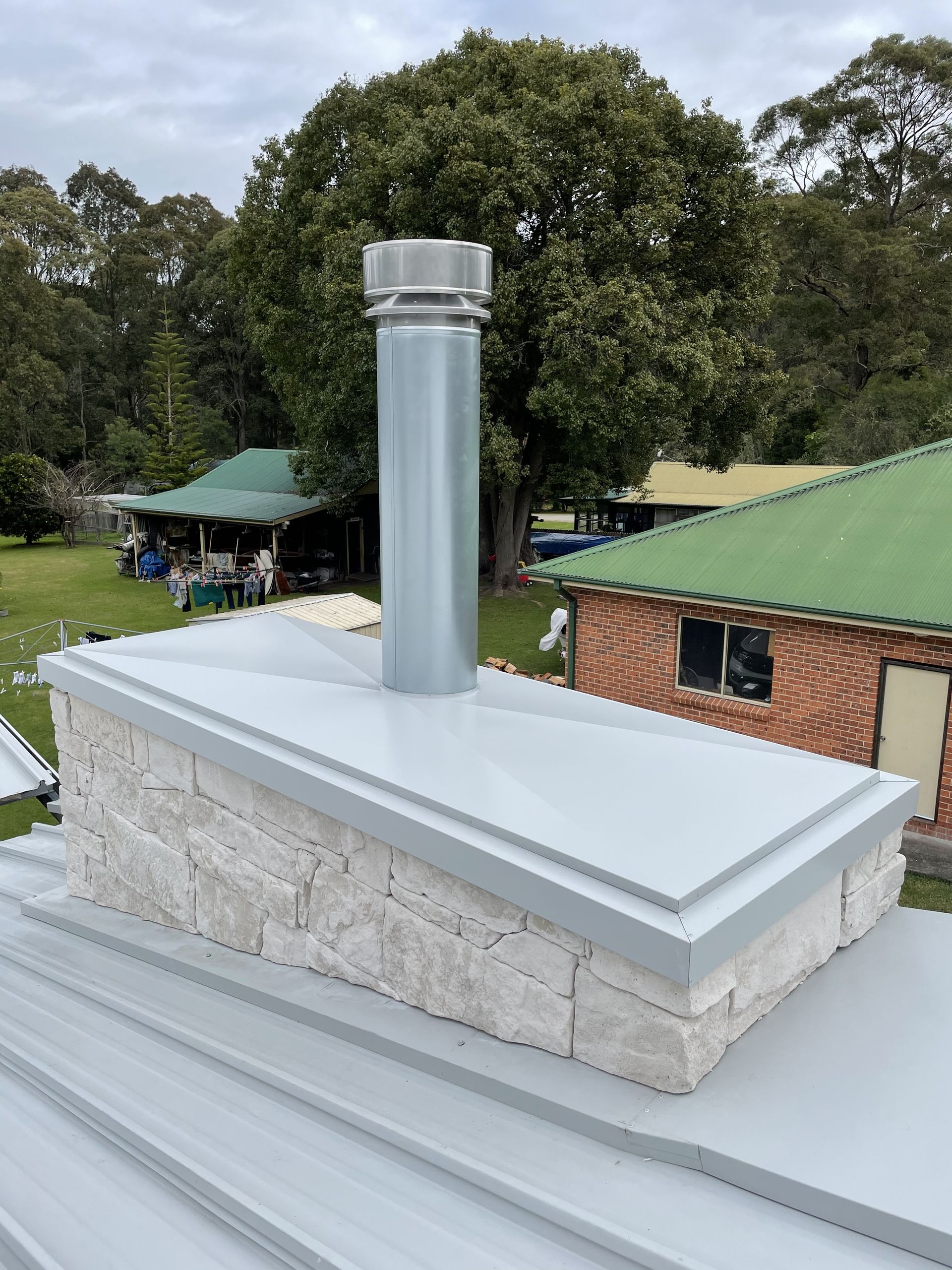 Chimney on a roof, constructed with stone and metal, topped with a silver flue. — R&M Sheetmetal Nowra Metal Roofing in South Nowra, NSW