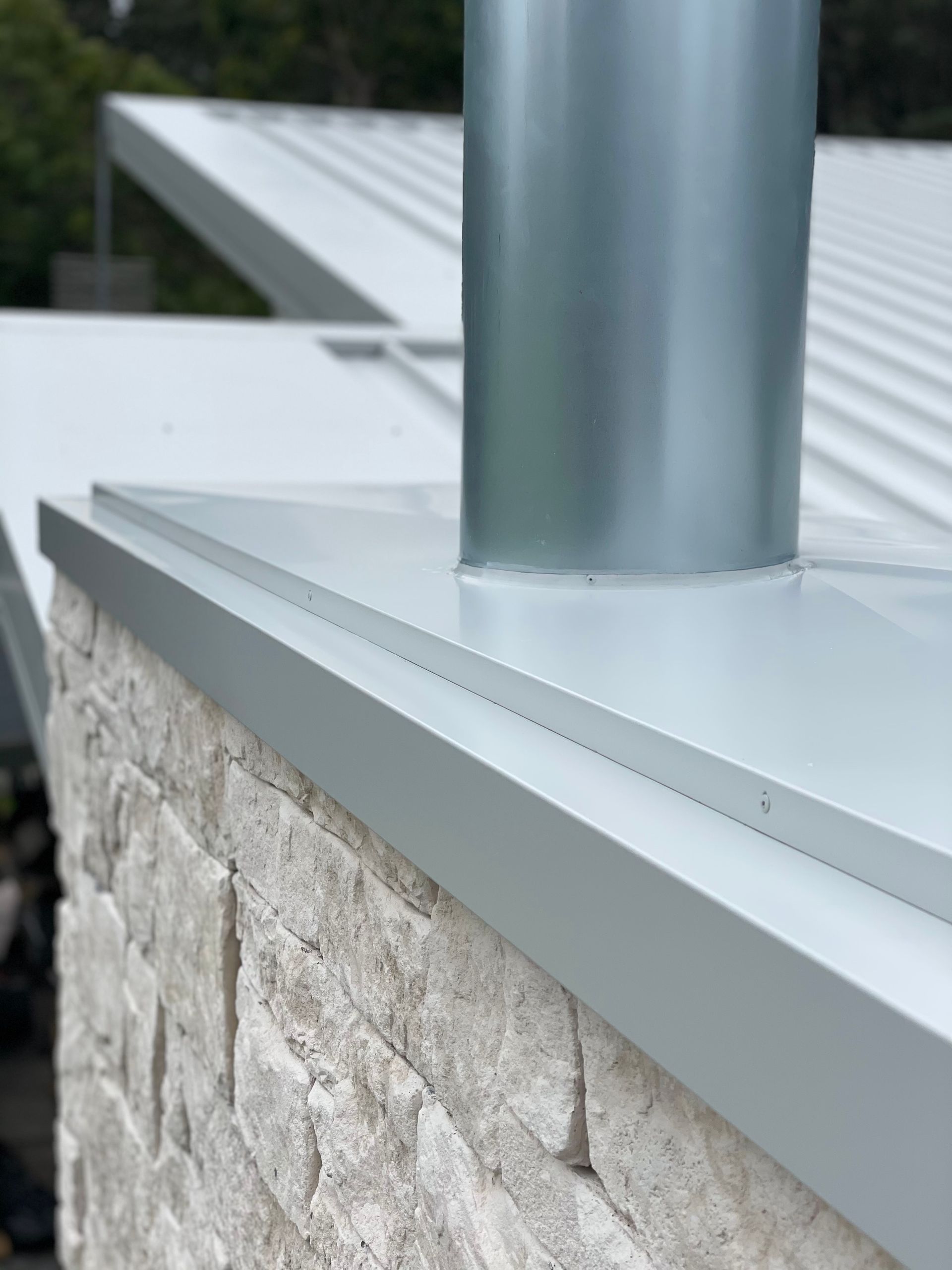 Close-up of a chimney on a white metal roof with light-colored stone wall. A silver, cylindrical chimney rises. — R&M Sheetmetal Nowra Metal Roofing in South Nowra, NSW