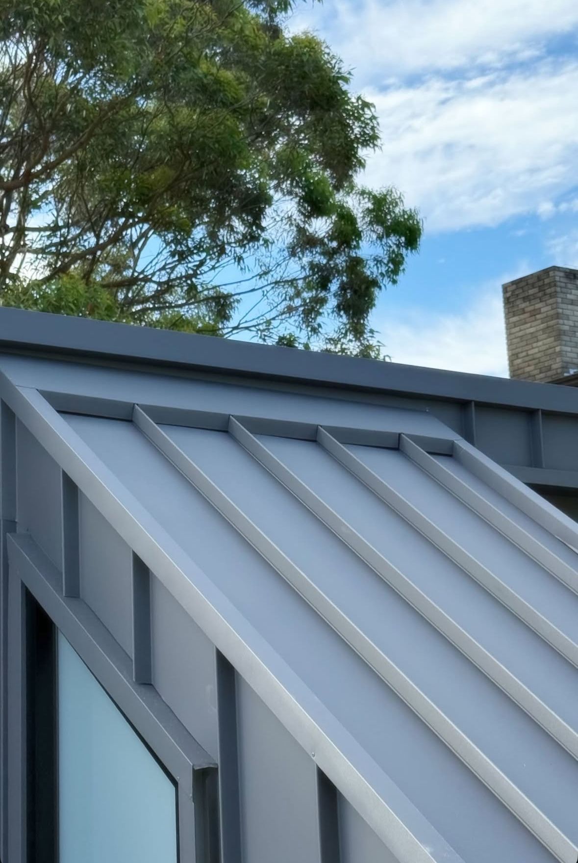 Gray metal roof with vertical ribs and a window frame against a blue sky, near a tree. — R&M Sheetmetal Nowra Metal Roofing in South Nowra, NSW
