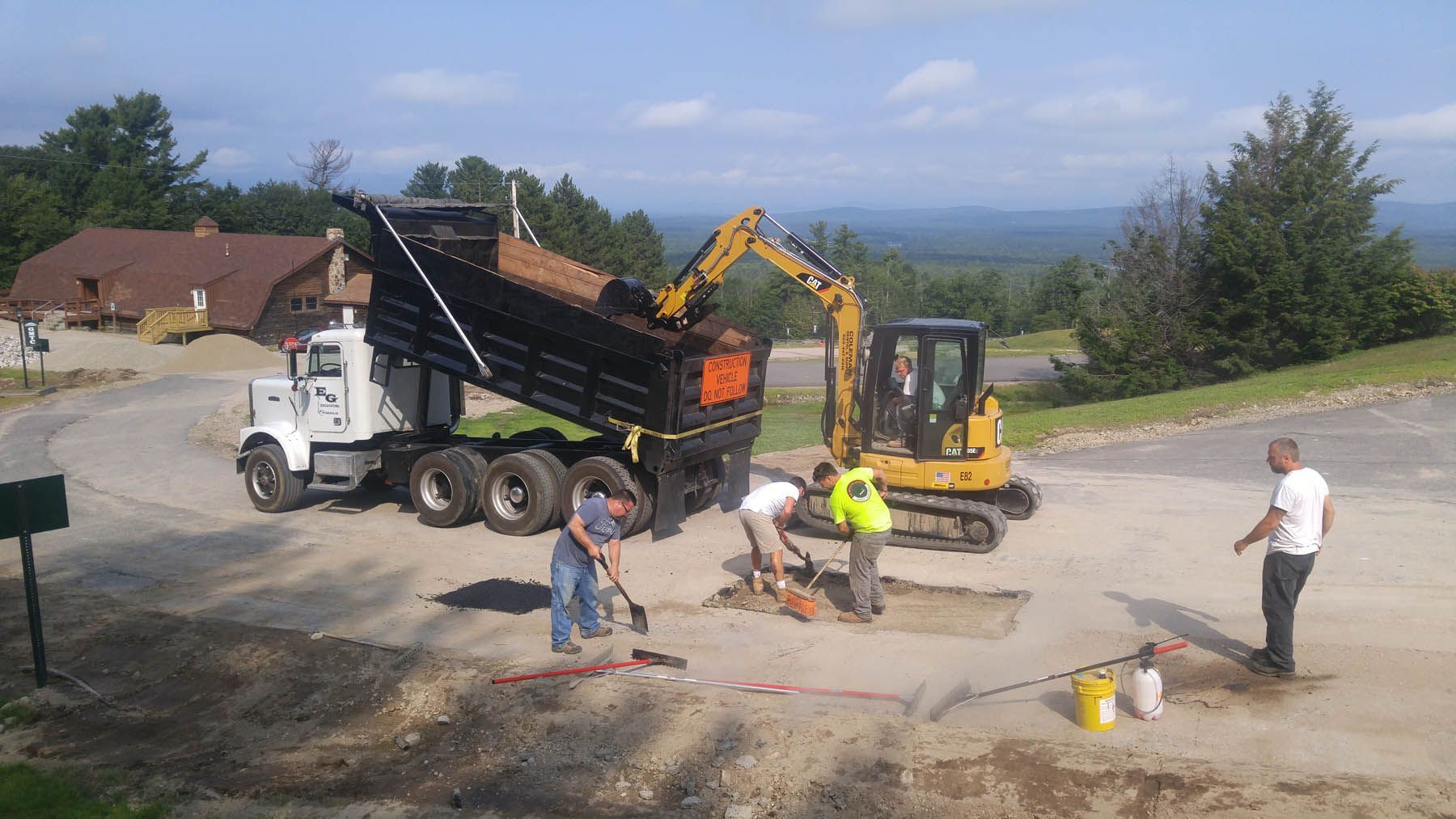 Construction site: Excavator loading dump truck with dirt, workers shoveling on a road, mountain backdrop.