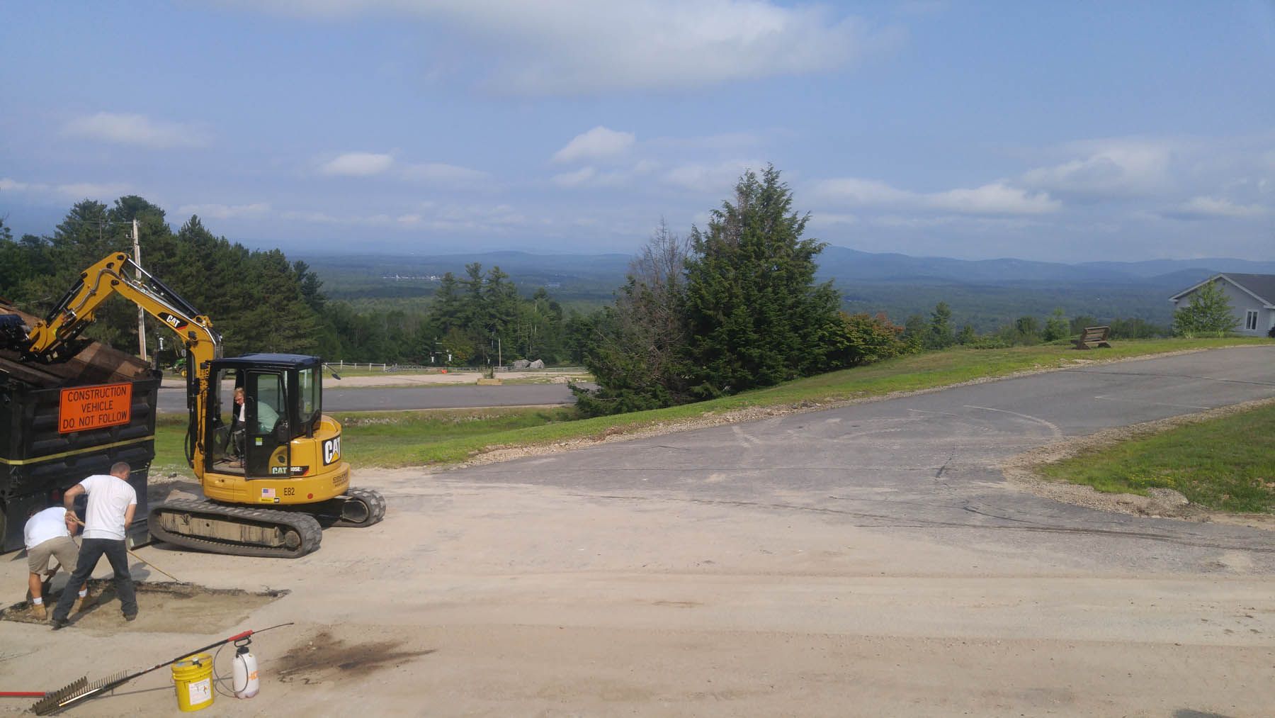 Construction site with a yellow excavator and two workers, overlooking a mountain range.