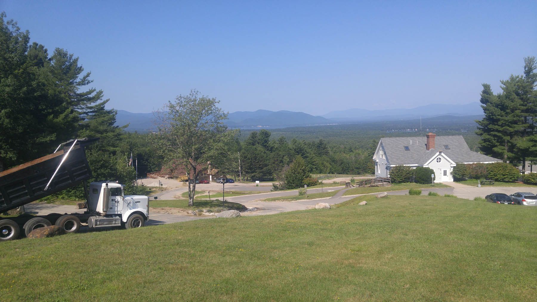 Truck unloading dirt onto a construction site near a white house, with a scenic mountain view.