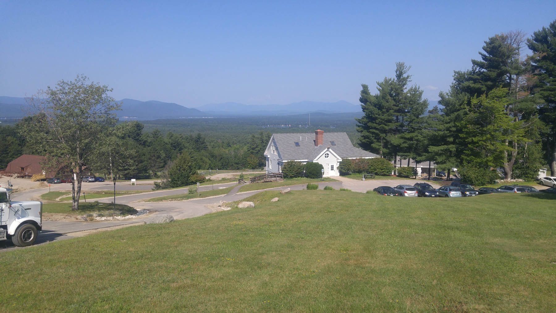 A white house on a grassy hill overlooking a forest and distant mountains under a clear blue sky.