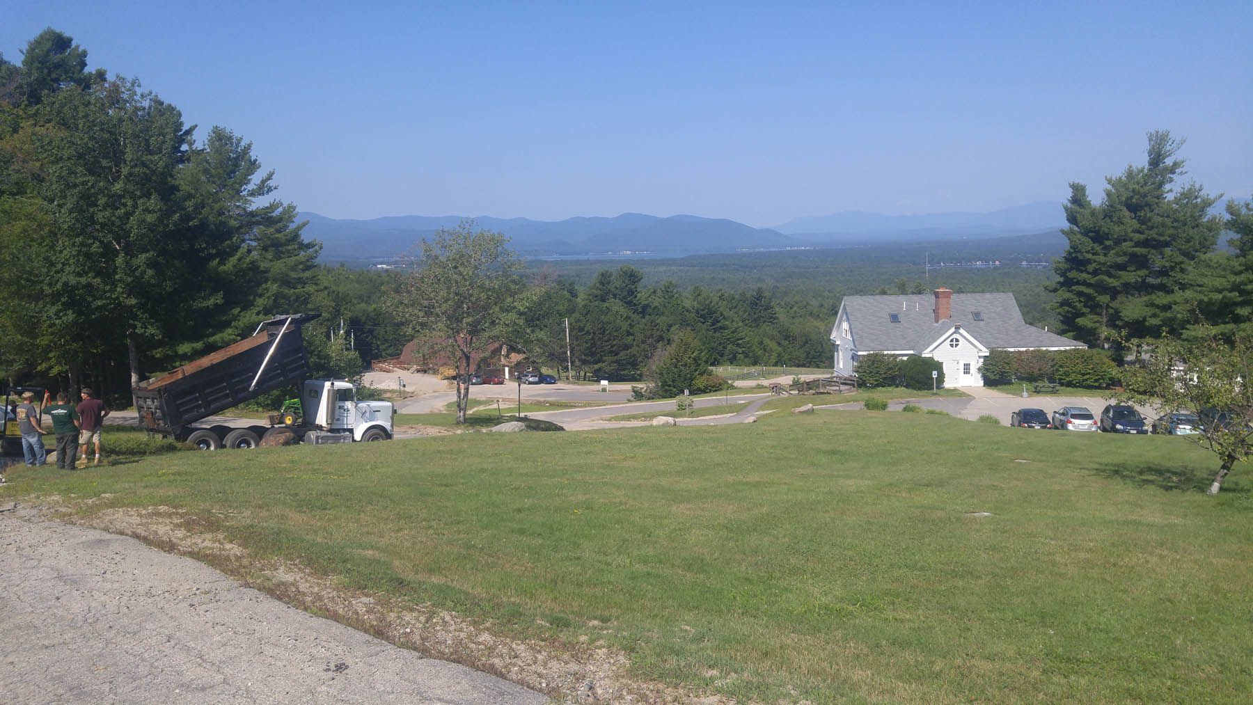 A house and other buildings on a grassy hill overlooking a distant valley under a clear blue sky.
