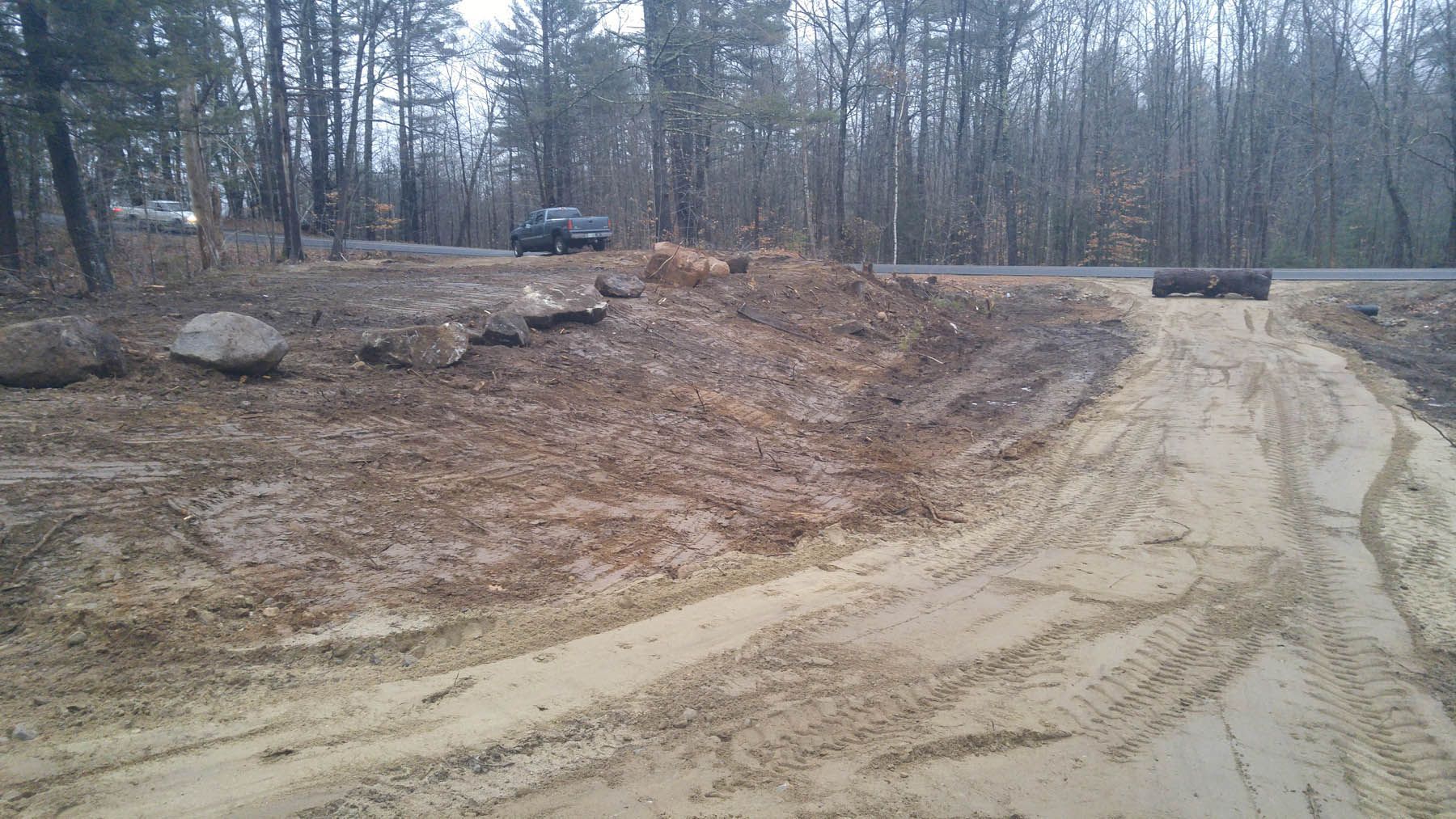 A dirt road leads uphill towards a road. Gray, muddy landscape with a few vehicles visible.