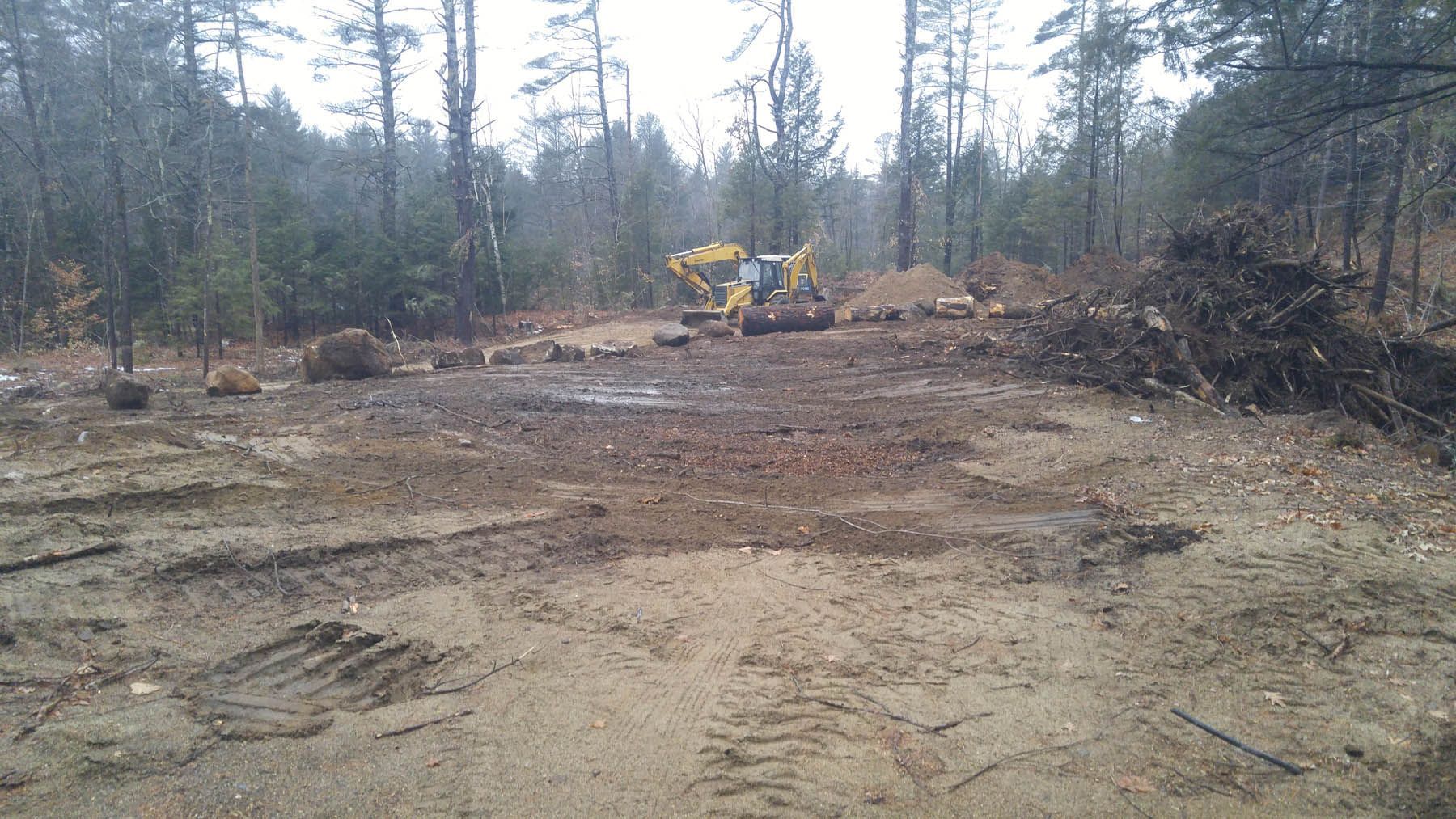 Construction site in a forest, excavator and cleared area, brown soil, trees in background.