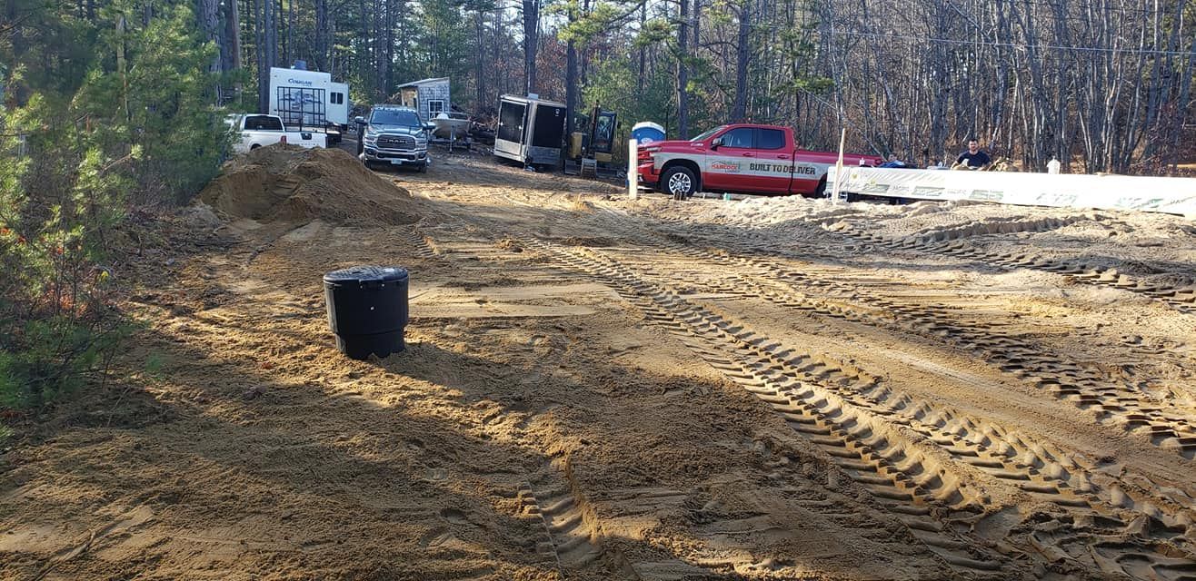 Dirt work site with trucks, RV, and equipment. A black barrel is in the foreground.