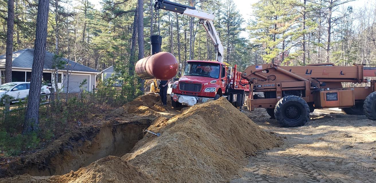 A crane lowering a red tank into a hole. A red truck is nearby with a house and trees in the background.