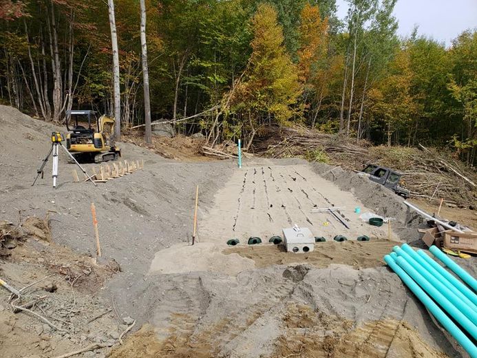 Construction site: Sand-filled area with piping. Small excavator and trees in the background.