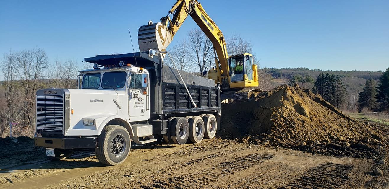 A yellow excavator loading a white dump truck with dirt in a rural setting on a sunny day.