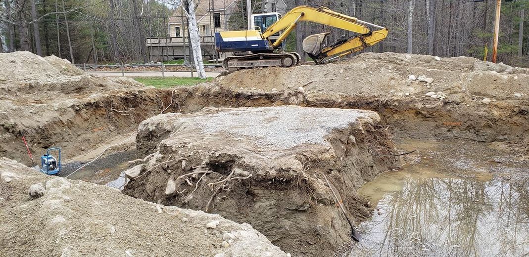 An excavator in a construction site, with a large excavated hole, partially filled with water.