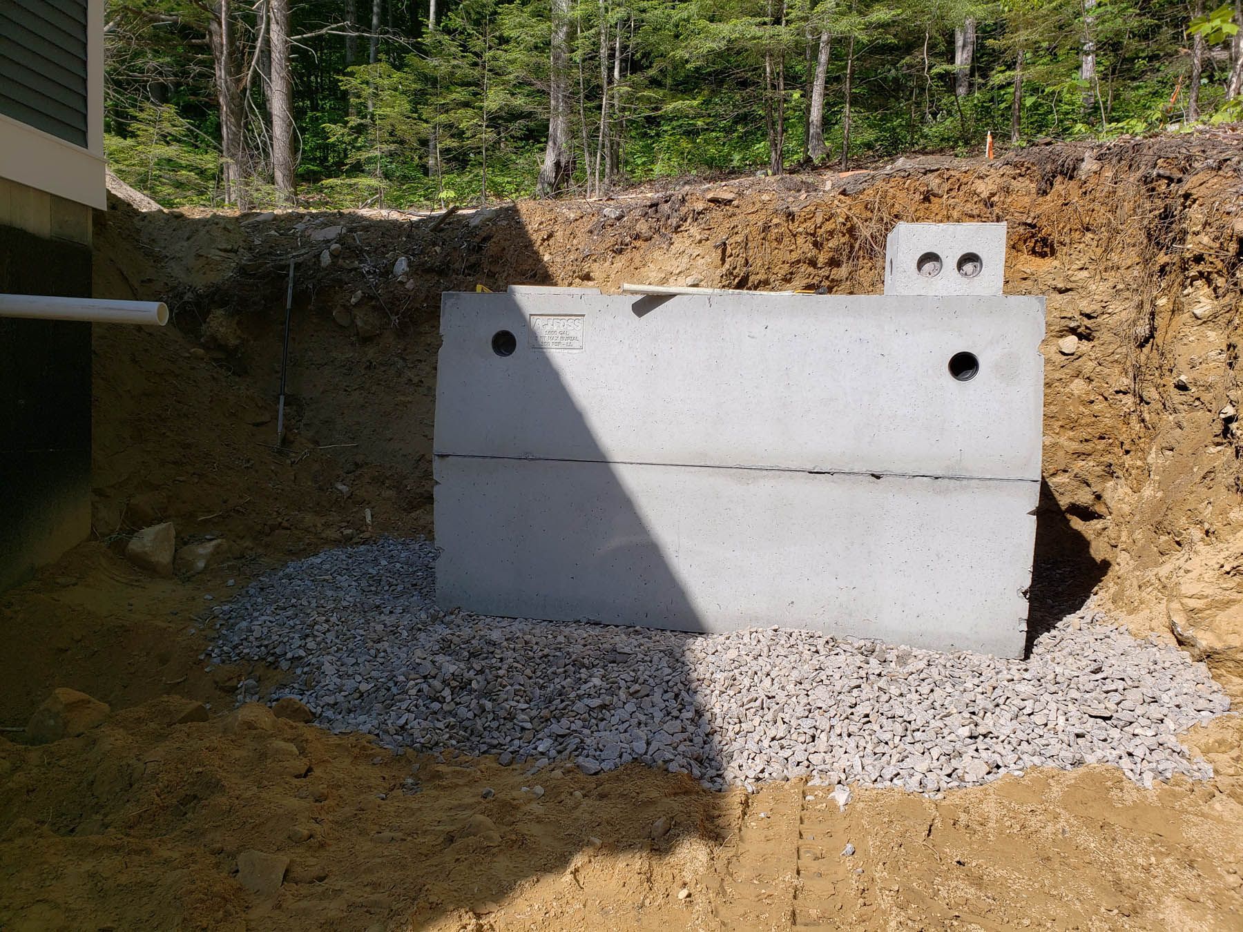 Concrete septic tank installed in a gravel-lined trench near a building, surrounded by dirt and a wooded background.