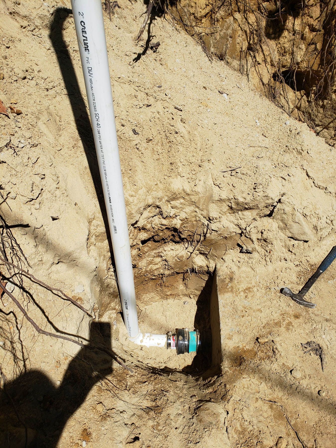 White PVC pipe in a sandy hole, connected to a dark fitting. A shovel handle is visible.